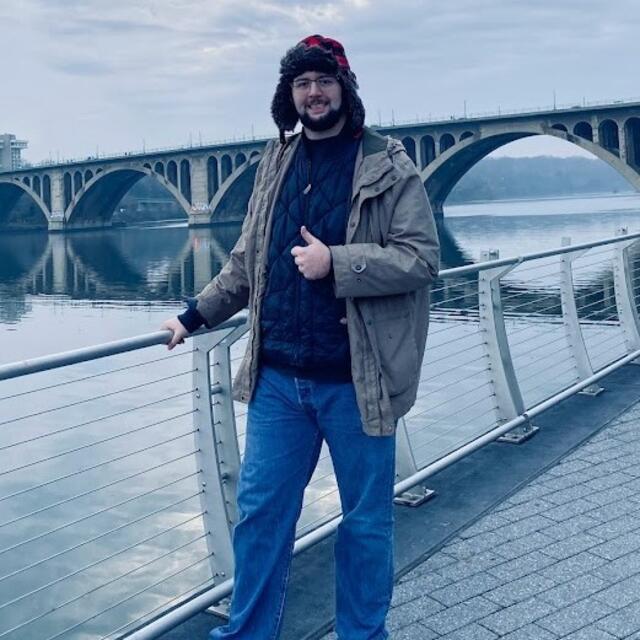 Large man wearing winter clothes in front of a river and bridge. He is giving a thumbs up.