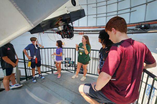 A university student in a green shirt explains the instruments of the telescope to several middle schoolers.