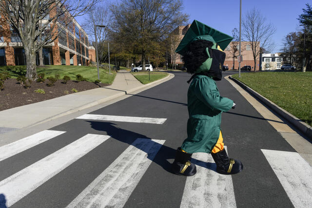 The Mason Patriot mascot walks across a road in a graduation gown and face mask.
