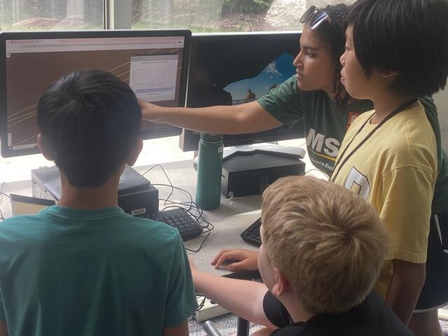 Flight Director Gabby is seen explaining the contents of a graph to three campers. All four individuals are looking at a computer screen showing a zoomed-in map of Mars.
