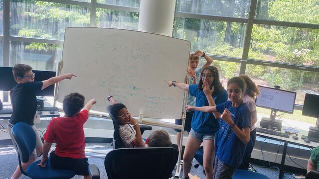 Several campers have gathered around a whiteboard, proud to show the photographer their work.