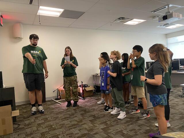 Several campers are assigning Ian instructions to write on the board. He appears to be struggling due to poor programming.