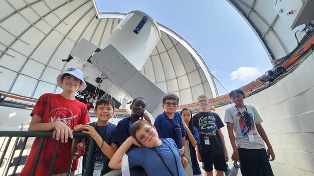 Many students are gathered the Geller Telscope. It is a sunny day as seen through the gap in the dome.