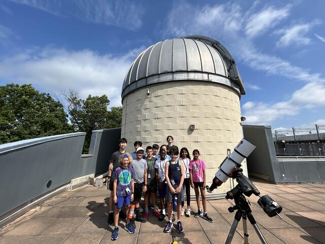 A group of students are gathered on the roof on a sunny day in front of the Observatory Dome and our Solar Telescope.