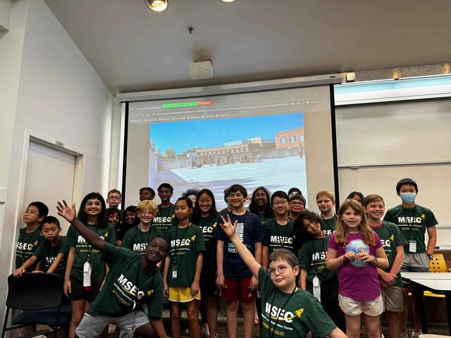 Many campers are gathered in a lecture hall. They are all wearing matching T-shirts. Two in front are doing jazz hands!