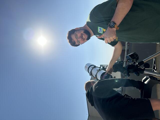 A Counselor and Camper are observing the Sun to spot the spots causing the recent aurora sightings. The counselor is looking to the camera and giving a thumbs up.