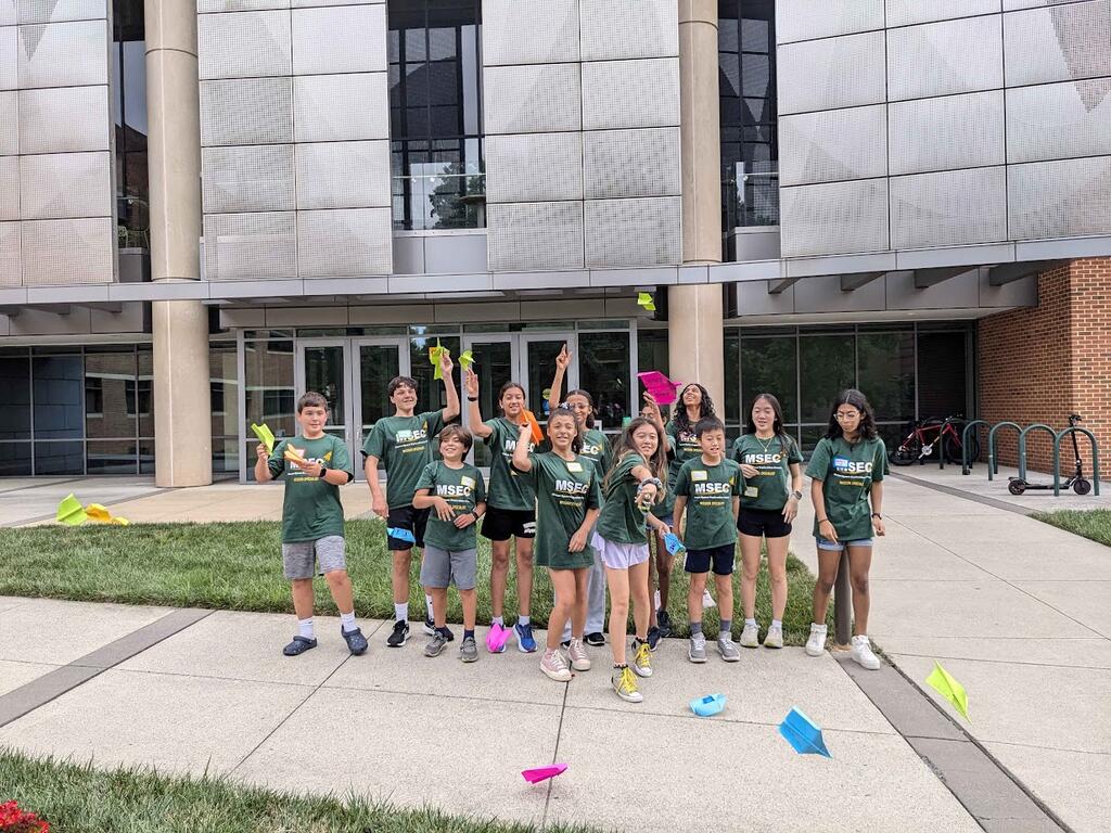 Several MSEC campers are gathered in front of Exploratory Hall. They are throwing colorful paper airplanes around the camera.