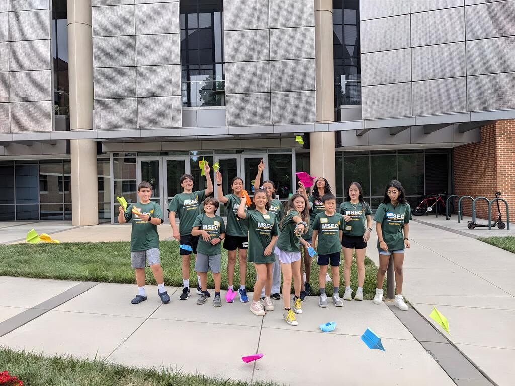 Many campers are situated in front of Exploratory Hall, throwing many colorful paper airplanes past the camera.